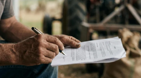 Gros plan sur des mains d'agriculteur tenant un stylo au-dessus de documents contractuels, avec un environnement agricole en arrière-plan